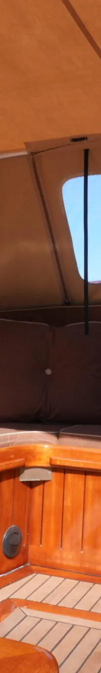 Een bootinterieur met bruine kussens, houten afwerking en een beige kap, met een vlag zichtbaar door een raam.