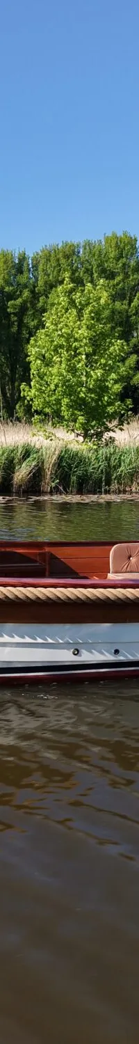 Een man vaart in een witte sloep met houten details en een Nederlandse vlag op een rivier met groene oevers.