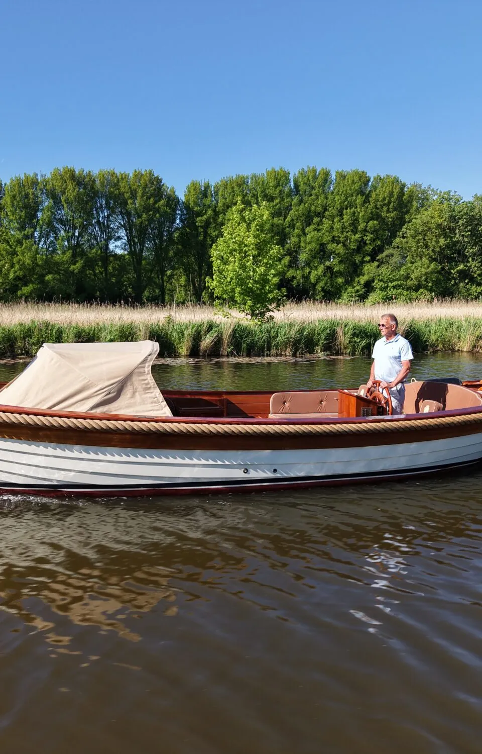 Een man vaart in een witte sloep met houten details en een Nederlandse vlag op een rivier met groene oevers.