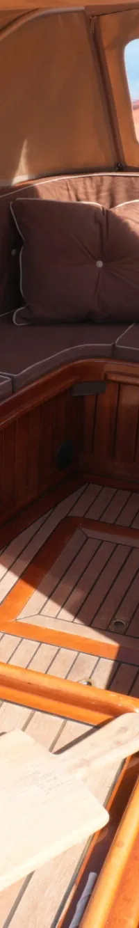 Een houten boot met bruine kussens en een beige kap ligt in het water, met andere boten op de achtergrond.