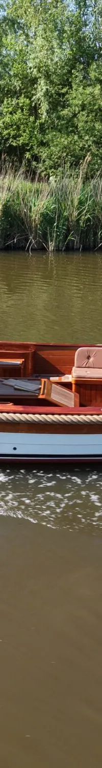 Een man staat in een houten sloep met een Nederlandse vlag aan de achtersteven, varend op een rivier met groene oevers.