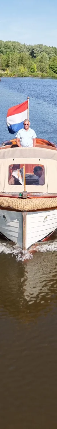 Een witte sloep met een Nederlandse vlag vaart over een rivier met een man aan het roer en twee passagiers.