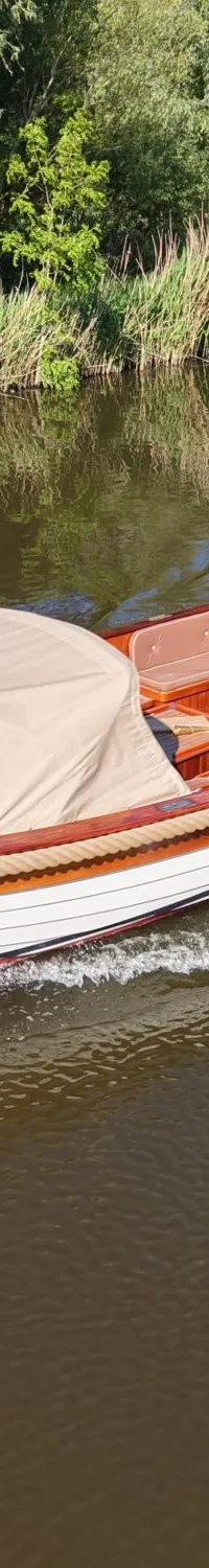 Een man vaart in een witte sloep met houten details en een beige kap op een rivier met groene oevers.