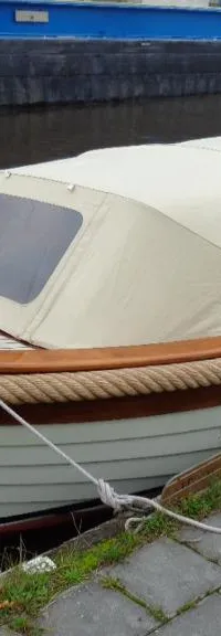Een witte boot met een beige kap en houten details ligt afgemeerd aan een kade met grijze tegels en gras.