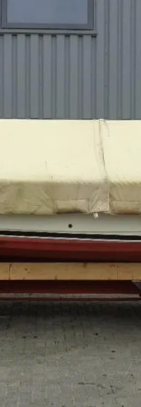 Een boot met een beige afdekzeil staat op een rode trailer met wielen voor een grijs gebouw met oranje deuren.