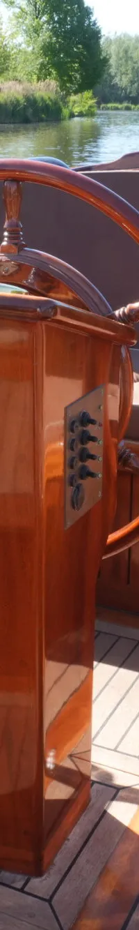 Een houten stuurwiel en een stuurstand met schakelaars zijn zichtbaar in een boot met bruine kussens en een houten dek.