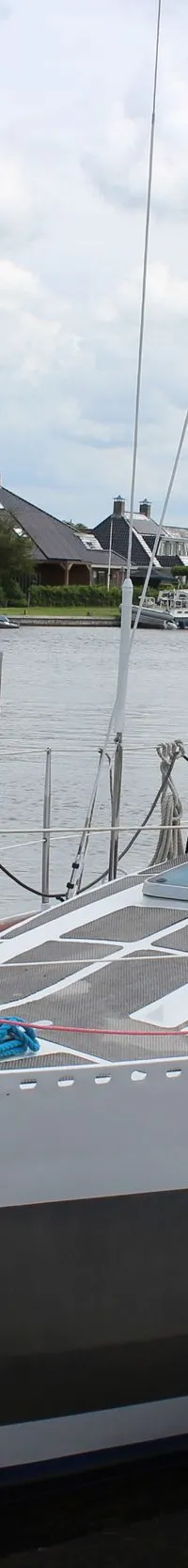 Een zeilboot met een lichtgrijze en donkergrijze romp ligt afgemeerd aan een houten steiger, met een "TE KOOP" bordje aan…