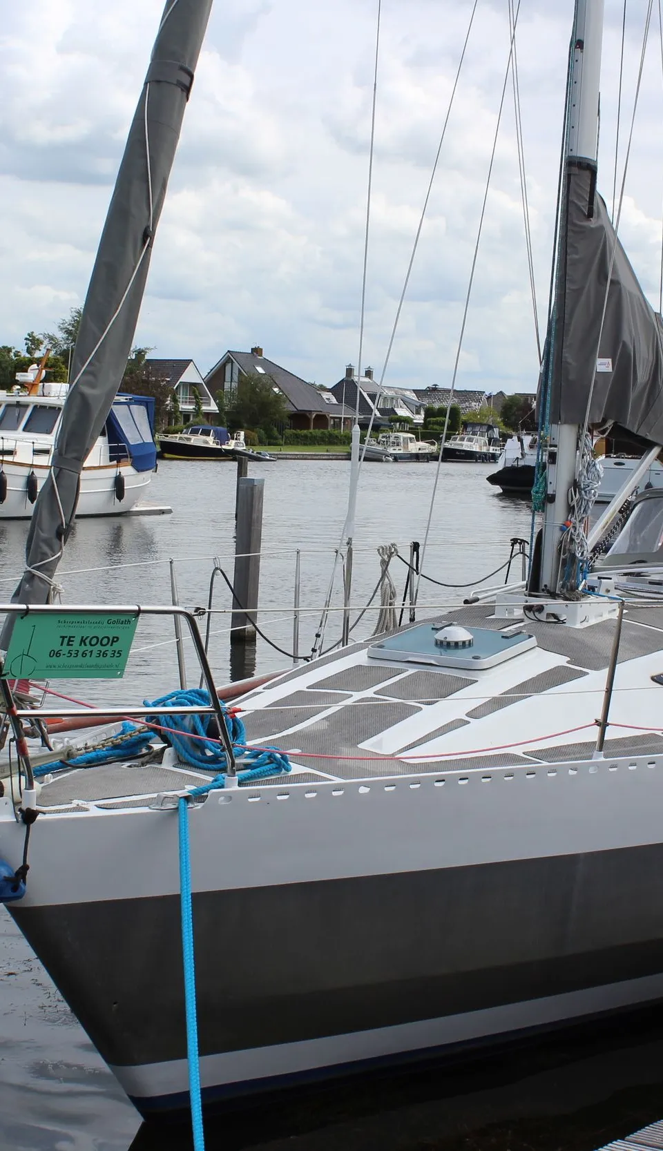 Een zeilboot met een lichtgrijze en donkergrijze romp ligt afgemeerd aan een houten steiger, met een "TE KOOP" bordje aan…