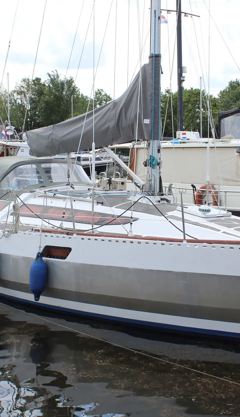 Een witte zeilboot met een grijze streep op de romp en een grijze zeilhoes ligt aangemeerd in een haven. — Ovni 32