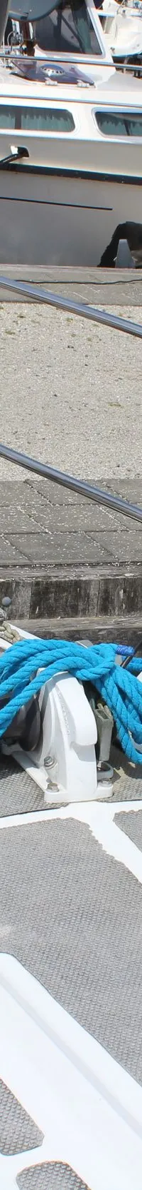 De boeg van een witte zeilboot met grijze antislipmatten ligt aangemeerd, met blauwe lijnen vast aan de houten kade.