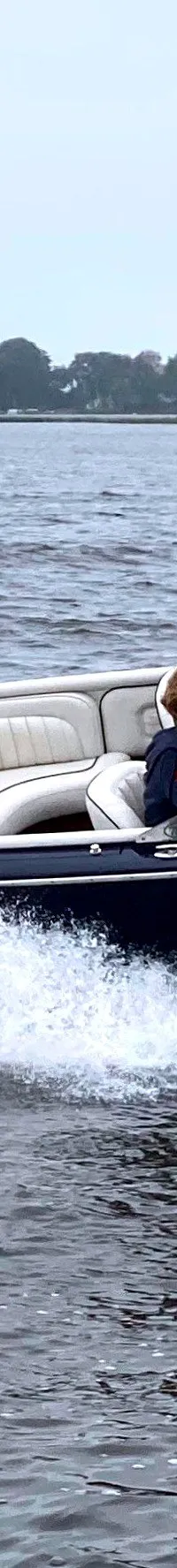 Een donkerblauwe speedboot met een houten dek en witte bekleding vaart over het water, met een persoon aan het stuur.