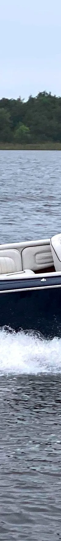 Een donkerblauwe speedboot met een blonde man aan het roer vaart over het water, met een bosrijke oever op de achtergrond.