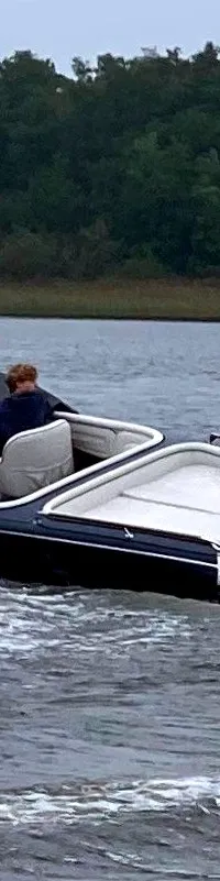 Een donkerblauwe motorboot met een houten dek en witte bekleding vaart op een meer met een bosrijke oever op de achtergrond.
