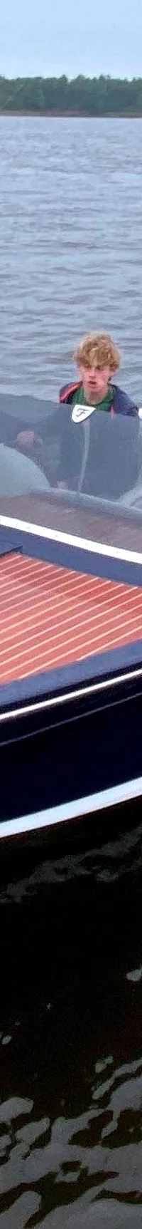 Een donkerblauwe speedboot met een houten dek en witte bekleding vaart op het water, met een persoon aan boord.