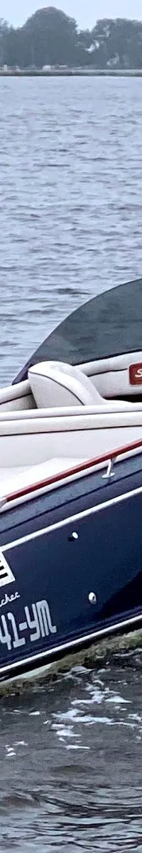 Een donkerblauwe motorboot met een witte romp en een houten dek vaart over het water met een persoon aan het roer.