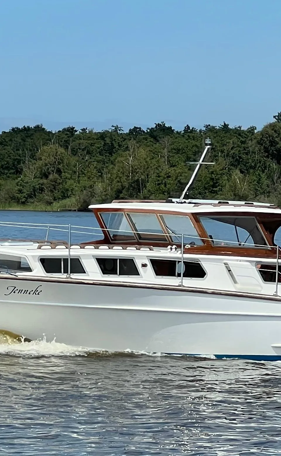 Een witte motorboot met de naam "Jenneke" vaart op het water, met een Nederlandse vlag aan de achterkant.