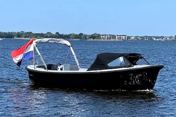Een zwarte boot met een Nederlandse vlag aan de achtersteven vaart op een meer met bebouwing op de achtergrond. — Asloep 770