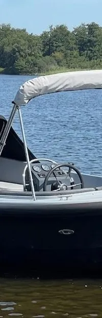 Een donkerblauwe sloep met een zwarte buiskap en een wit zonnedek ligt in het water, met een Nederlandse vlag aan de…