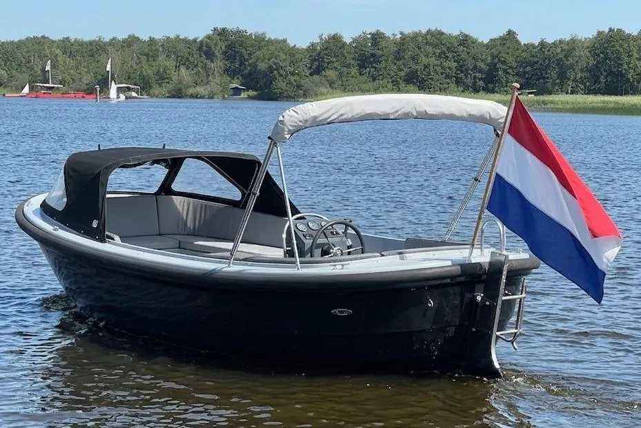 Een donkerblauwe sloep met een zwarte buiskap en een wit zonnedek ligt in het water, met een Nederlandse vlag aan de…