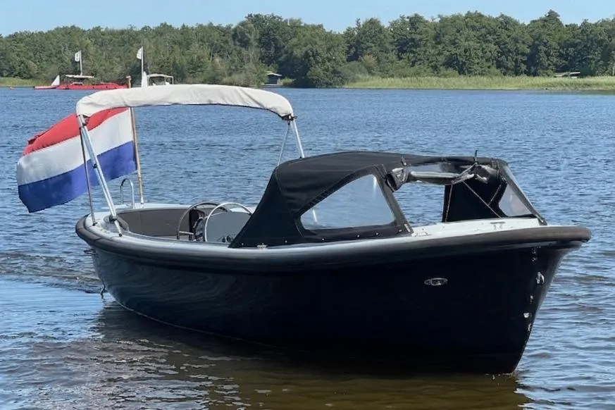 Een donkerblauwe boot met een Nederlandse vlag, een zwarte buiskap en een witte biminitop ligt in het water. — Asloep 770