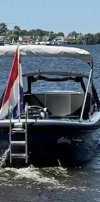 Een donkergrijze boot met een Nederlandse vlag en een zwemtrap vaart op een meer met een beboste oever op de achtergrond.